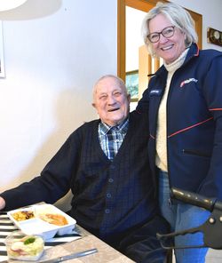 Alter Mann sitzend neben stehender Frau, beide lächeln.