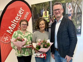 Zwei Frauen und ein Mann stehen nebeneinander vor einer Malteser Fahne. Die Frauen haben einen Blumenstrauß in der Hand.