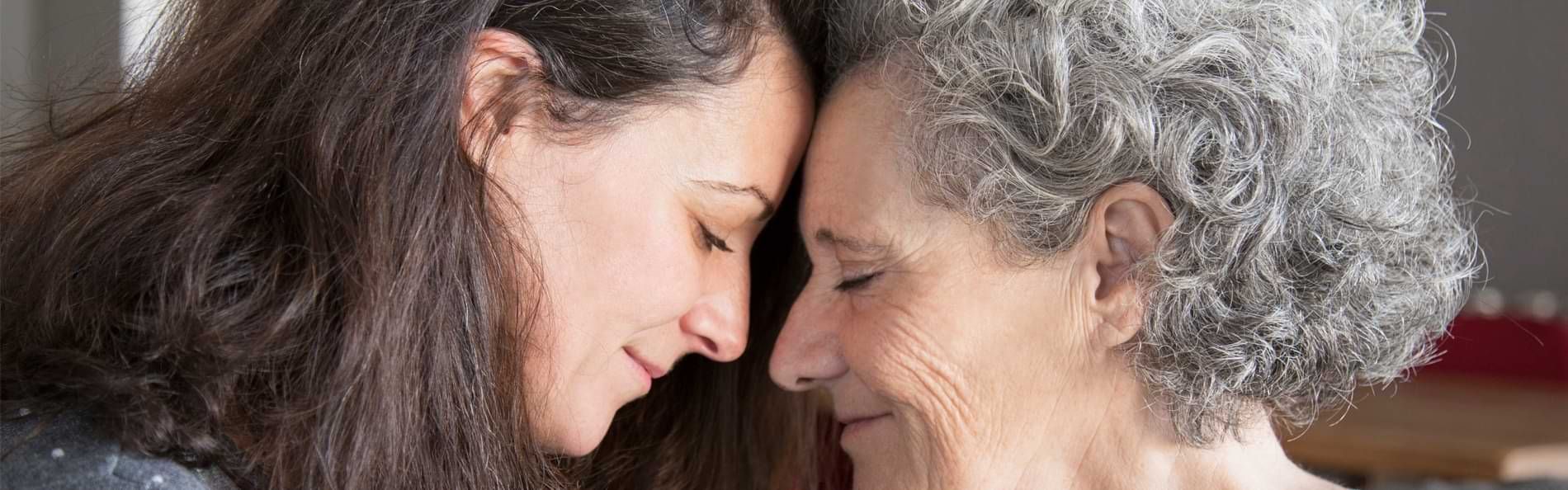 Eine junge Frau und eine ältere Frau lächeln und legen mit geschlossenen Augen ihre Köpfe aneinander. 