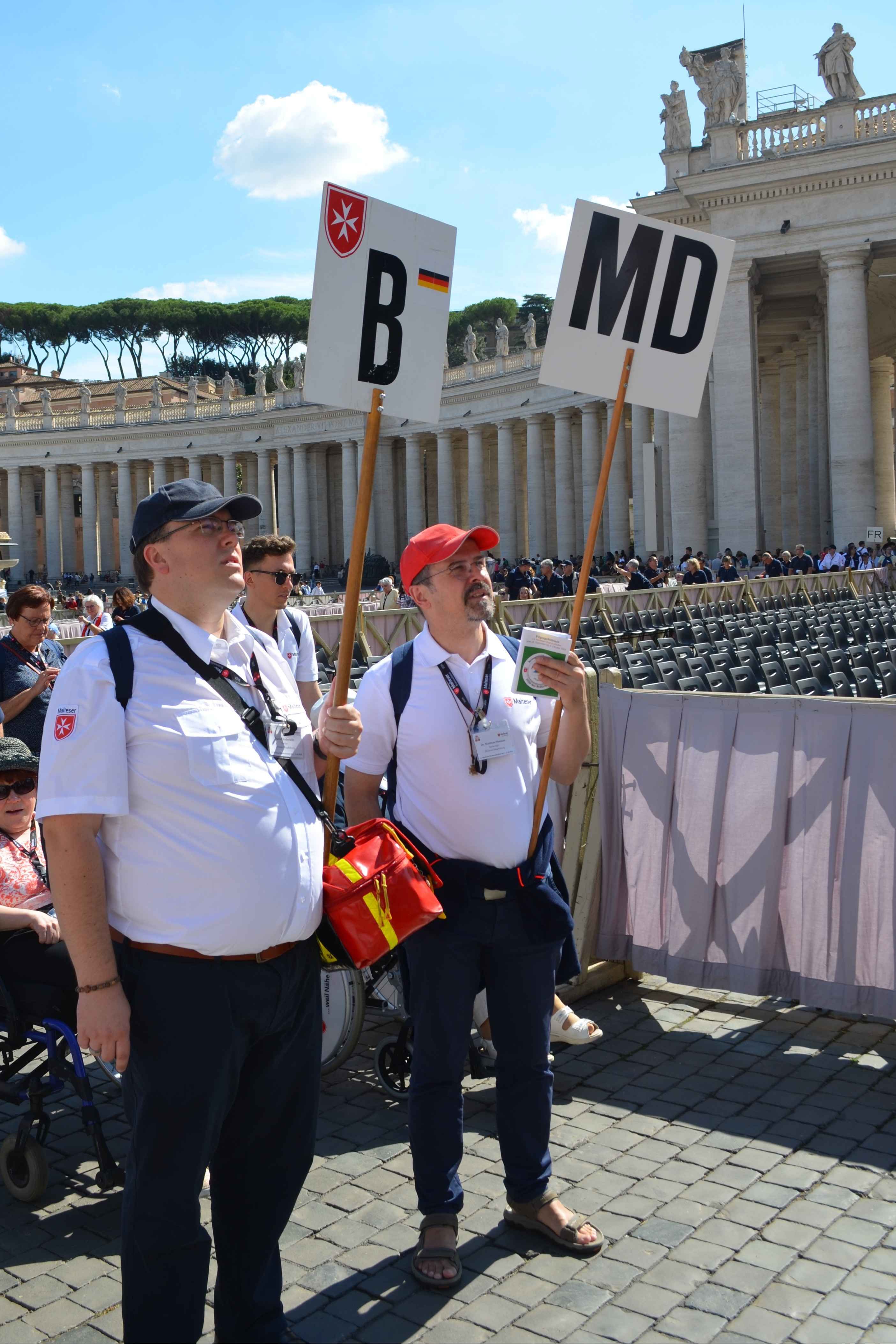 Zwei Männer in weißen Poloshirts und roten Mützen halten Schilder mit den Buchstaben 'B' und 'MD' vor einer historischen Säulenfassade.