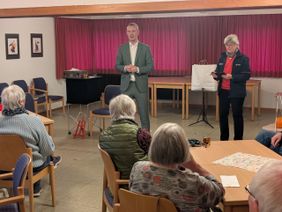 Eine weitere freiwillige Ehrenamtliche der Malteser steht rechts neben Jan Phillip Lehmker und soll sich ein Wort ausdenken und uneinsehbar aufschreiben.