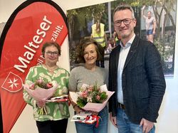 Zwei Frauen und ein Mann stehen nebeneinander vor einer Malteser Fahne. Die Frauen haben einen Blumenstrauß in der Hand.