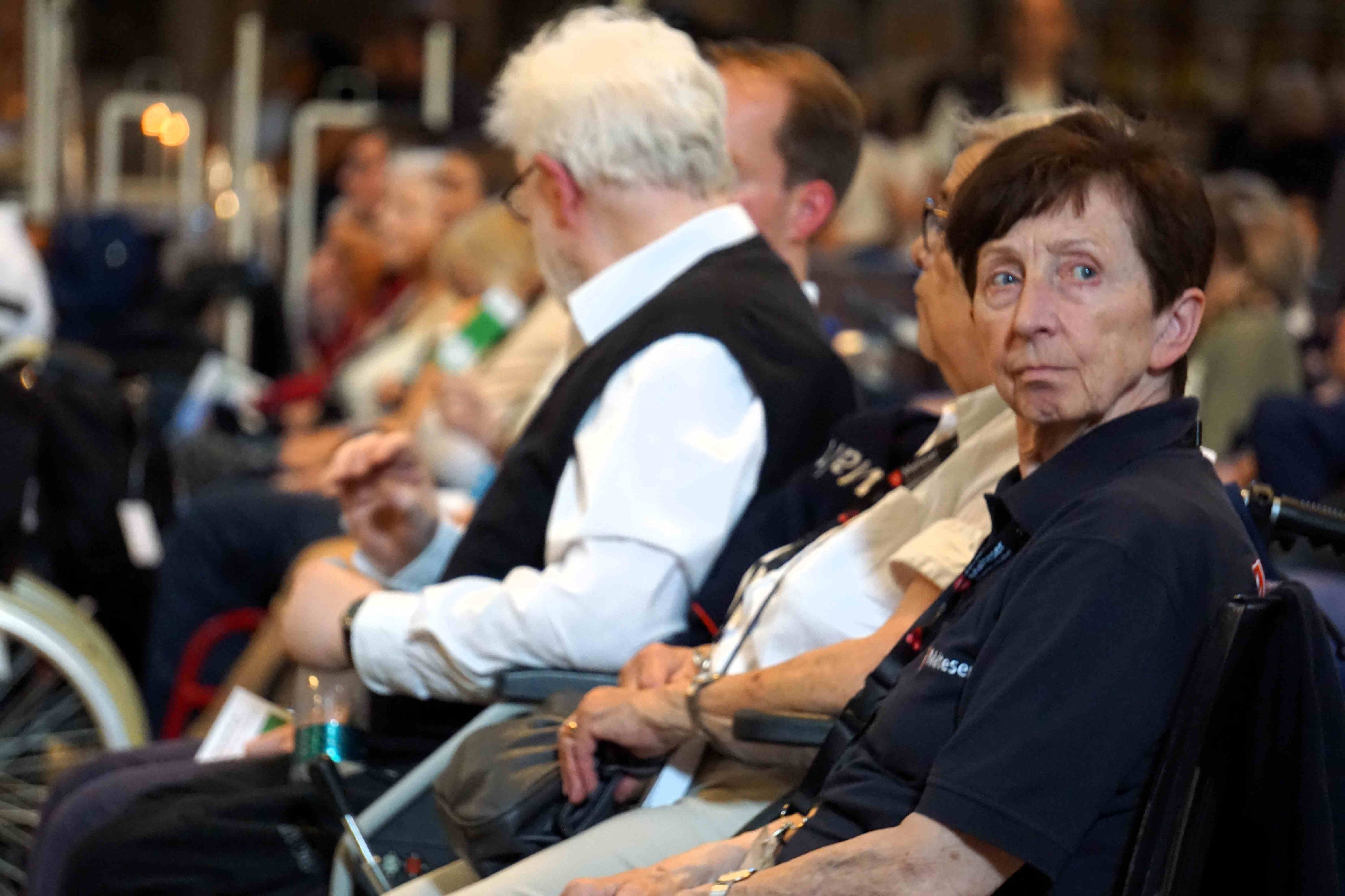 Mehrere Personen sitzen in einer Reihe, einige in Rollstühlen, in einem Innenraum einer Kirche mit unscharfem Hintergrund.