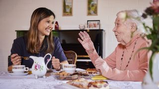 Junge Ehrenamtlerin des Besuchsdienstes sitzt mit einer Seniorin bei Kaffee und Kuchen zusammen.