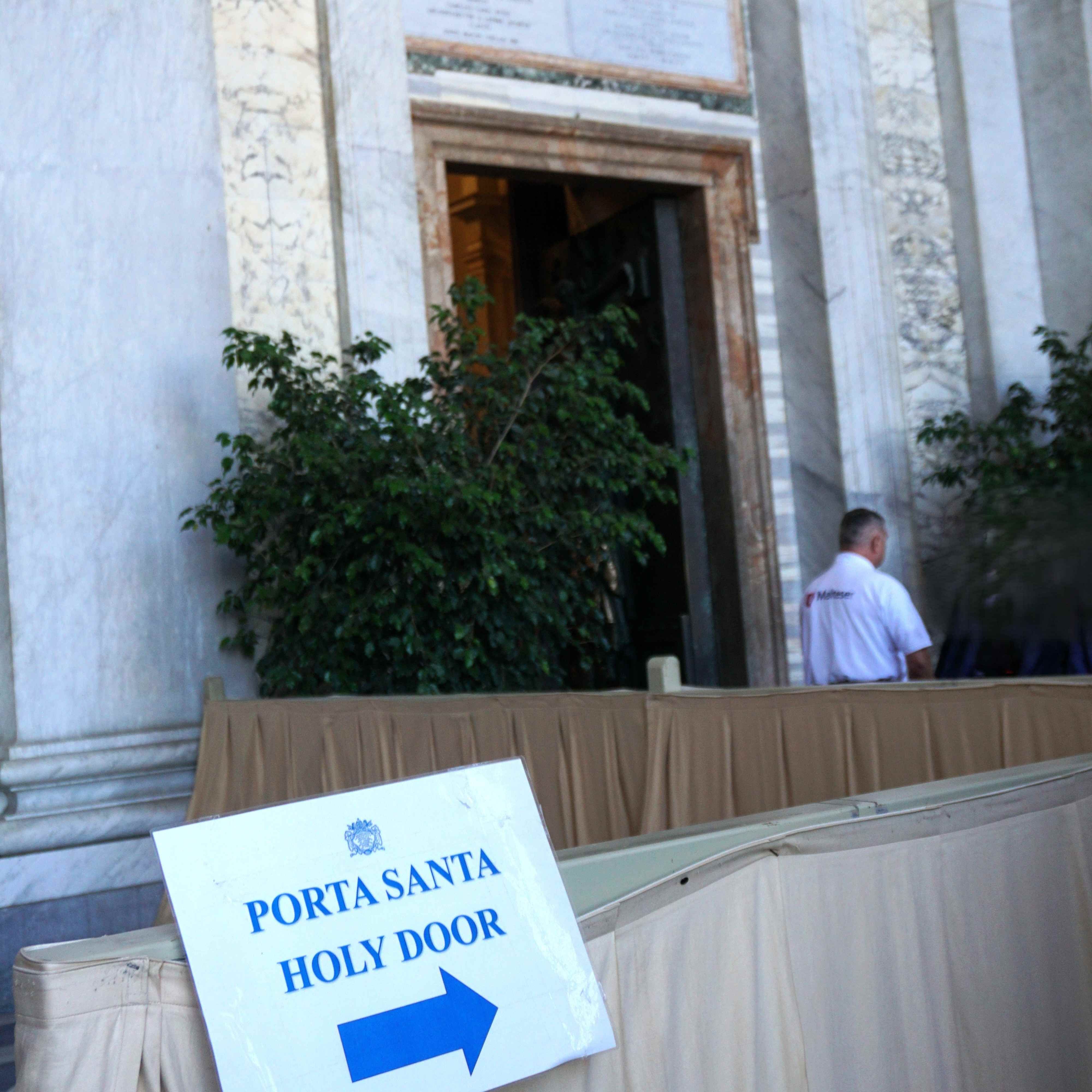 Eingang der Heiligen Pforte Lateran mit offenem Tor, rechts ein Mann in weißem Hemd, davor Schild mit blauem Pfeil und Text 'PORTA SANTA HOLY DOOR'.