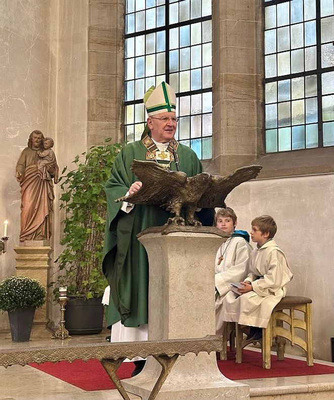 Ein Bischof steht in einer Kirche am Pult, während zwei Messdiener neben ihm sitzen.