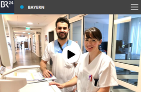 Zwei Pflegekräfte an einem Stationsarbeitsplatz mit Dokumenten und Computern in einem hellen Flur.