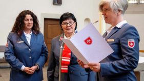 Drei Frauen sind zu sehen. Eine liest aus einem Umschlag.