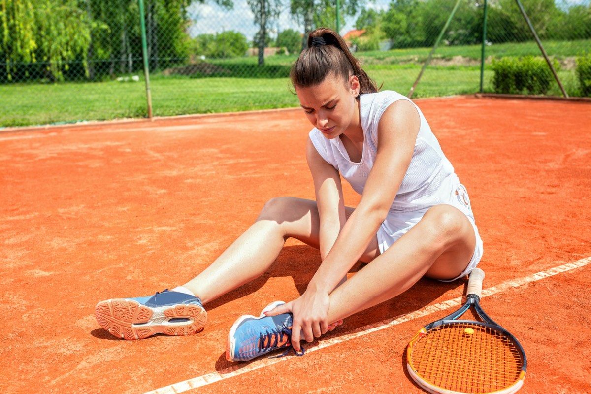 Junge Frau auf einem Tennisplatz hält sich den Fußknöchel mit schmerzverzerrtem Gesicht