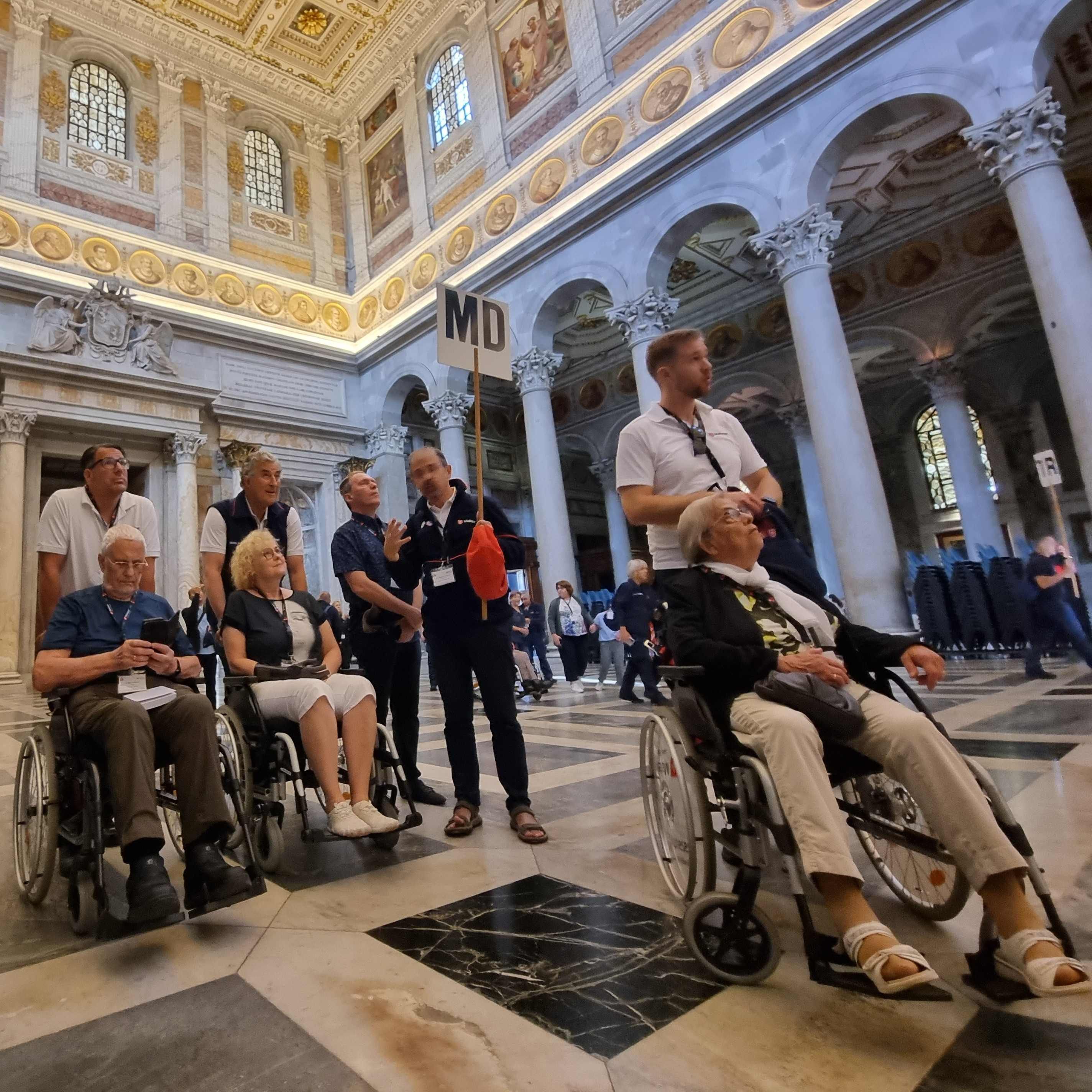Menschen in Rollstühlen und Begleitpersonen in einem prunkvollen Kirche mit hohen Säulen und verzierten Wänden.