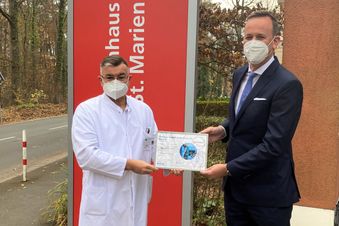 Arzt und Mann im Anzug stehen vor einem Malteser Schild und halten gemeinsam eine Urkunde.