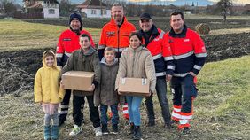 Vier Männer in Malteserkleidung stehen hinter vier Kindern und Jugendlichen, von denen zwei ein Hilfspaket in der Hand halten.