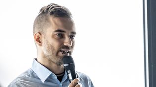 Person hält Vortrag mit Mikrofon und steht vor heller Präsentationsfläche.