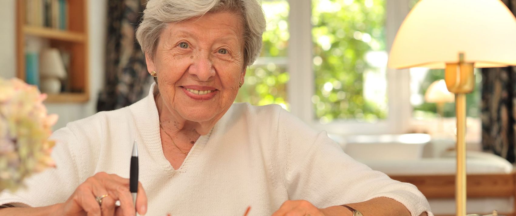 Eine ältere Frau sitzt mit Stift und Zettel an einem Schreibtisch