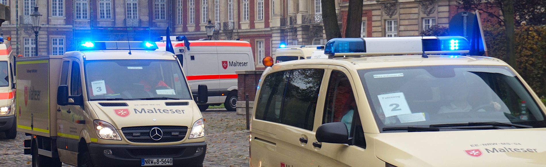 Malteser Einsatzfahrzeuge fahren mit eigeschaltetem Blaulicht auf die Kamera zu. Im Hintergrund das münsteraner Schloss.