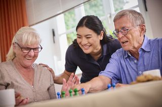 Drei Personen sitzen gemeinsam an einem Tisch vor einem Fenster und spielen ein Brettspiel mit bunten Spielfiguren. Eine Person im Hintergrund beugt sich unterstützend über die Spielenden, während Tassen und Spielmaterial auf dem Tisch stehen.