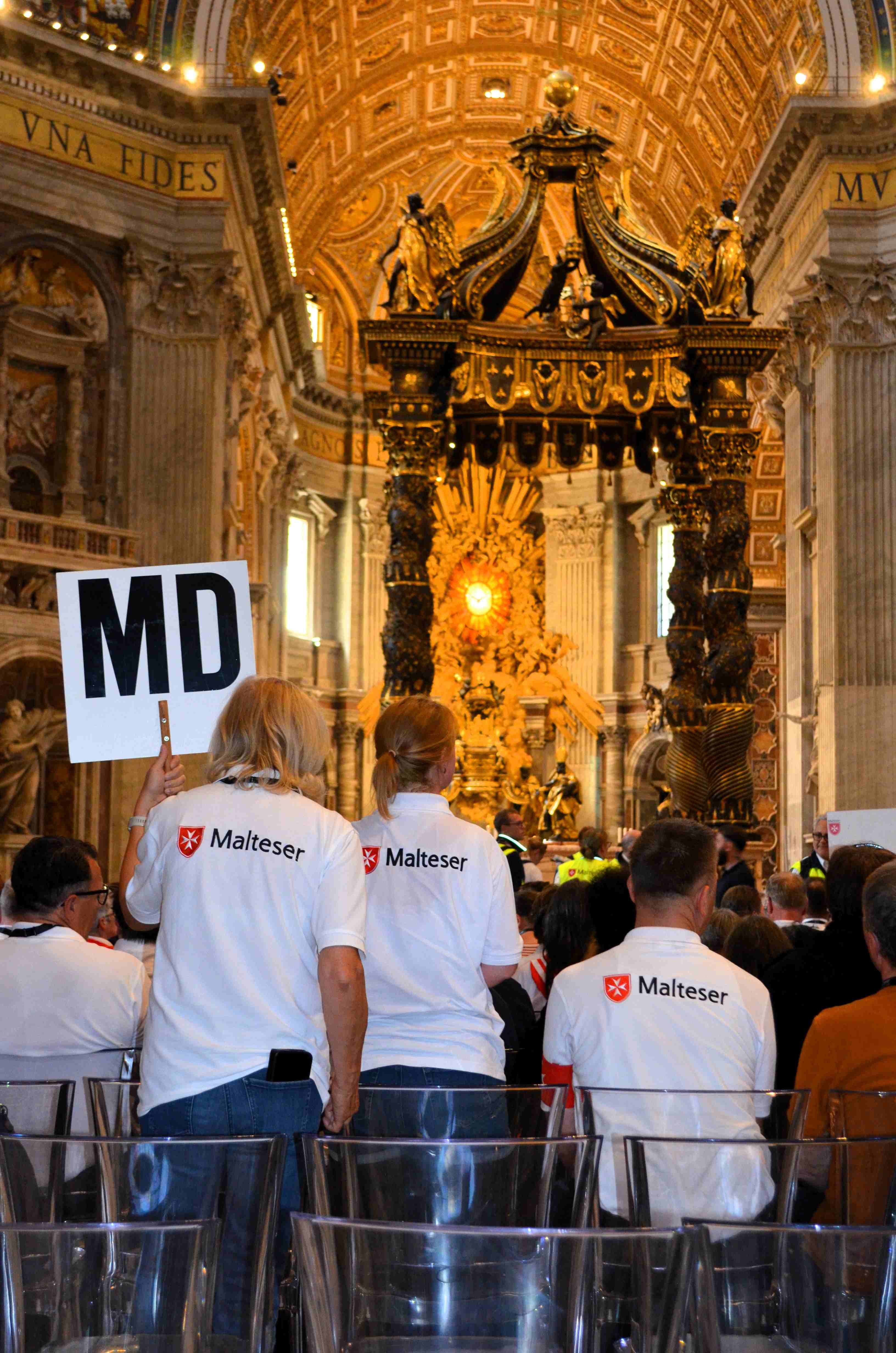 Menschen mit weißen T-Shirts und rotem Schriftzug stehen in einer Kirche, eine Person hält ein Schild mit den Buchstaben 'MD' hoch.