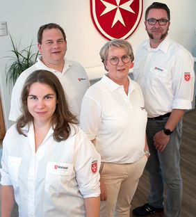 Zwei Frauen und zwei Männer stehen vor dem Malteserwappen.