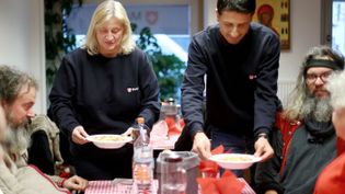 Blonde Frau und schwarzhaariger Mann in Malteser Pullis servieren zwei bärtigen Männern Teller mit Suppe 