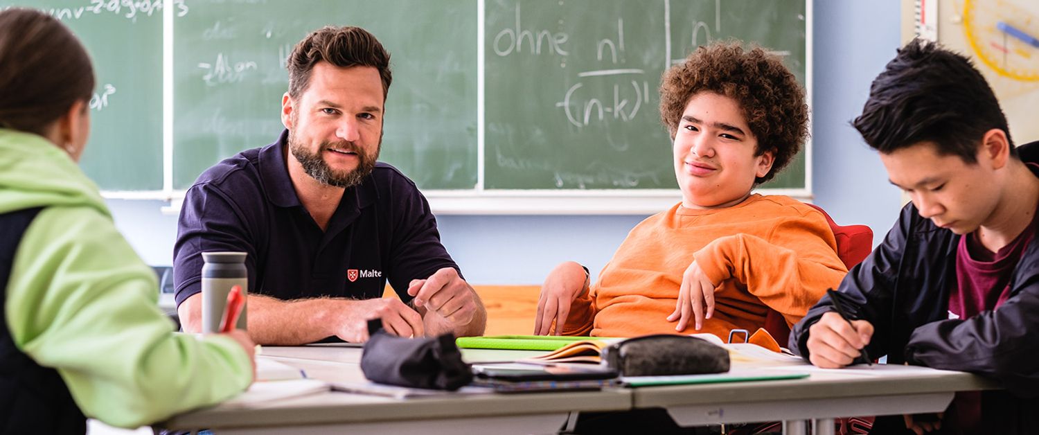 Malteser Schulbegleiter sitzt mit seinem Schüler und anderen Schülern in der Klasse. 