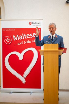 Ein Mann in Malteser-Uniform spricht an einem Rednerpult vor einem Malteser-Banner mit Herzsymbol.