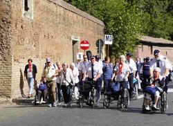 Gruppe von Menschen, darunter mehrere im Rollstuhl, geht eine Straße entlang neben einer hohen Steinmauer mit Verkehrsschildern, darunter ein rotes Einfahrtverbotsschild.