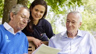 Zwei ältere Männer und eine junge Frau sprechen über ein Dokument, dass einer der Männer in den Händen hält