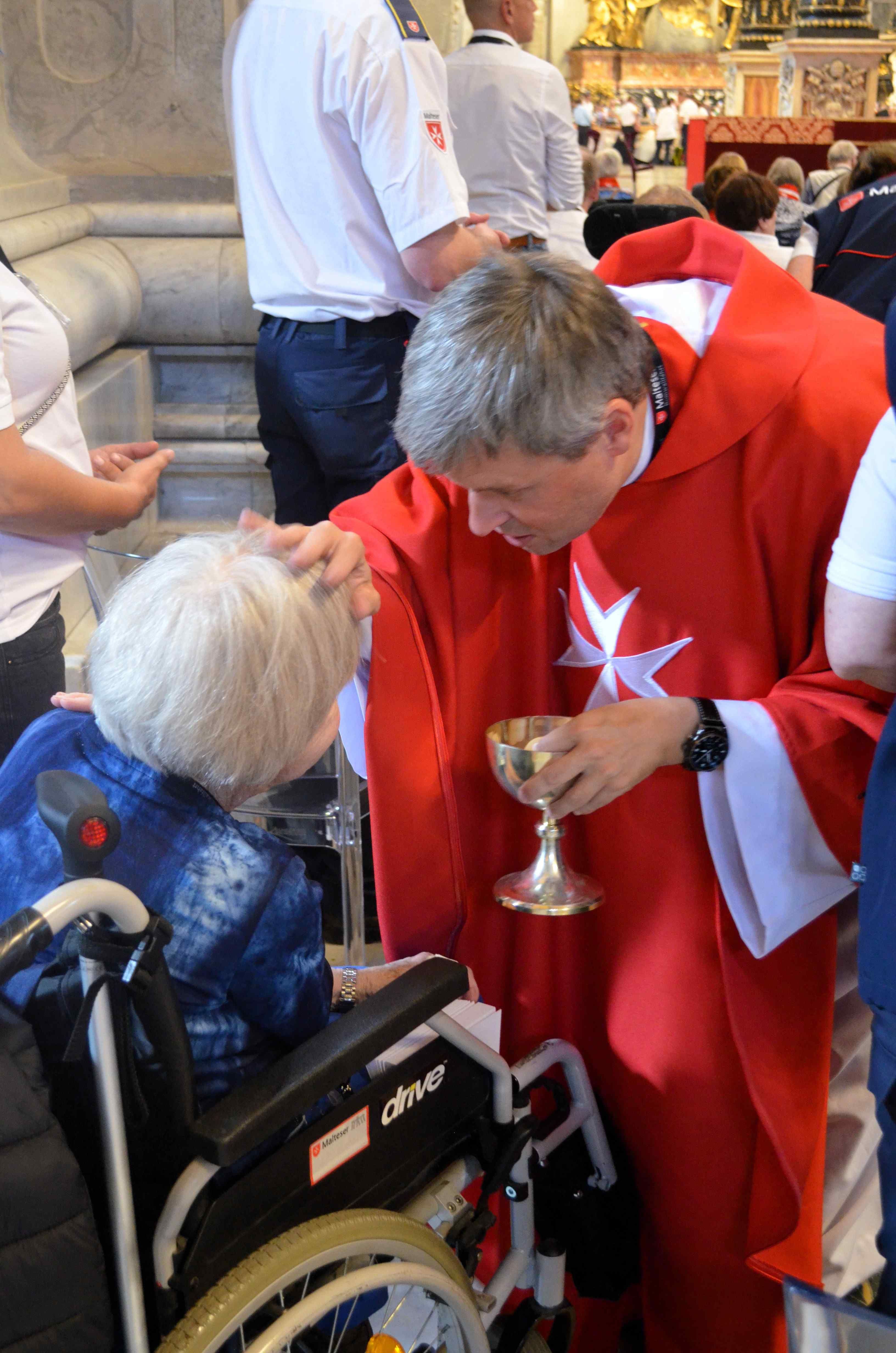 Priester in rotem Gewand hält Kelch und segnet sitzende ältere Person im Rollstuhl in einer Kirche.