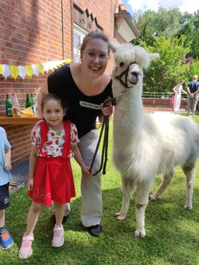 Ein Kind und eine erwachsene Frau stehen gemeinsam mit einem Alpaka im Garten und lächeln