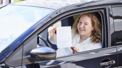 Eine junge Frau sitzt hinter dem Lenkrad eines PKW und hält eine Bescheinigung durch das geöffnete Fenster.