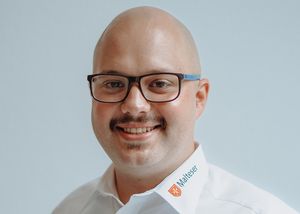 Ein 30 Jahre alter Mann lächelt in die Kamera. Er trägt ein Hemd mit Malteser-Beschriftung.