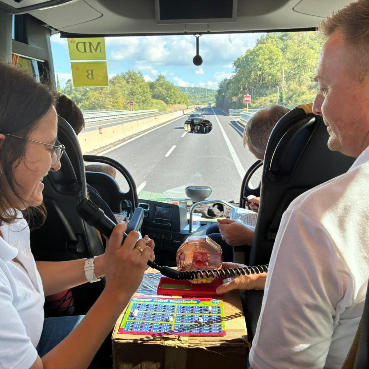 Blick aus dem Busfahrerbereich auf die Autobahn, eine Person hält ein Mikrofon, auf dem Armaturenbrett liegt ein Spielplan mit bunten Zahlen.