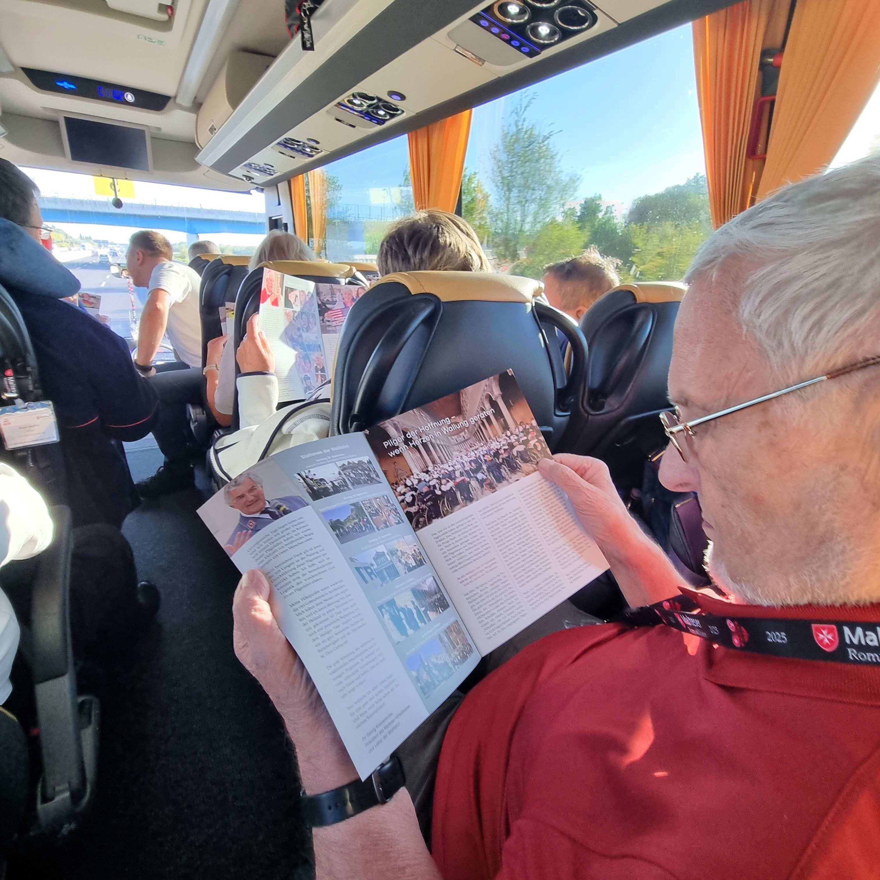 Person in rotem Oberteil liest eine mehrseitige Broschüre im Bus mit orangefarbenen Vorhängen und mehreren sitzenden Fahrgästen.