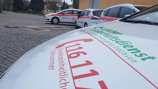 Drei weiße Autos mit roter und grüner Aufschrift 'Ärztlicher Bereitschaftsdienst' und Notrufnummer 116117, geparkt auf Kopfsteinpflaster vor einem Gebäude.
