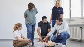 Ein Mann kniet neben einer Ausbildungspuppe und zeigt auf einen Punkt des Oberkörpers, eine Malteser Ausbilderin und weitere Personen sitzen und stehen um ihn herum.