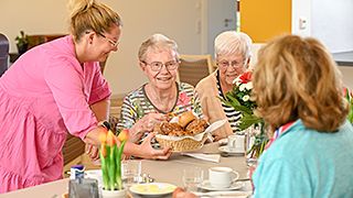 Eine Frau reicht Brötchen älteren Damen die am Tisch sitzen