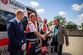 Mehrere Männer begrüßen sich. Hinter ihnen steht ein Krankenwagen für Herzenswünsche.