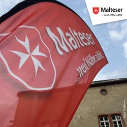 Auf dem Foto ist eine Beachflag in Rot mit dem Logo der Malteser und dem Schriftzug Malteser weil Nähe zählt vor einem Gebäude im Hintergrund zu erkennen.