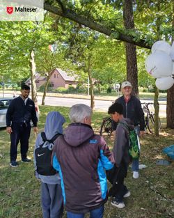 Mehrere Besucherinnen und Besucher stehen auf einer Grünfläche unter schattigen Bäumen am Malteser-Stand und nehmen an einem Spielangebot teil. Rechts im Bild sind weiße Malteser-Luftballons zu sehen, im Hintergrund eine Straße sowie Fahrräder.
