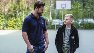 Ein Mann in dunkelblauem Poloshirt und Jeans steht auf einem Sportplatz neben einem Jungen mit schwarzer Jacke und grauem T-Shirt, beide mit Händen in den Taschen.
