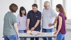 Ein Malteser Ausbilder zeigt vier umstehenden Personen an einer Kinder-Übungspuppe das Überstrecken des Kopfes..