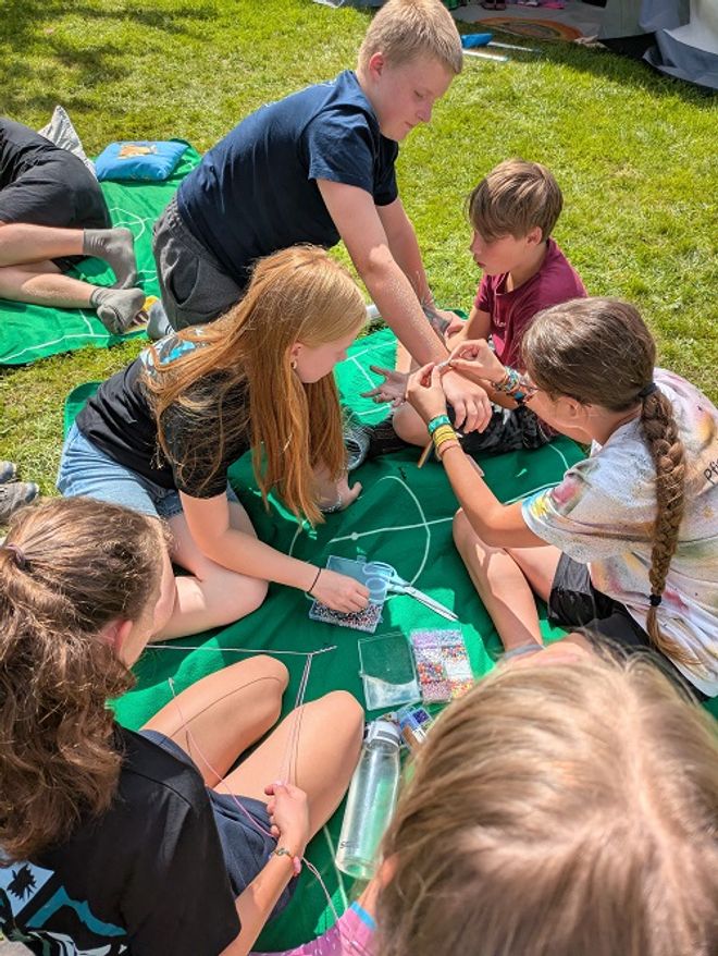 Mehrere Kinder und Jugendliche, die auf einer Decke im Gras sitzen und Armbänder gestalten