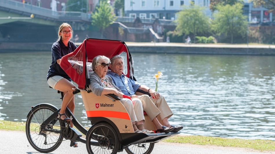 Älteres Paar sitzt in einer Rikscha, die von einer Frau gefahren wird, im Hintergrund ein Fluss.