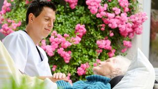 Ein junger Pfleger sitzt am Bett einer alten Frau. Das Bett steht auf der Terrasse.