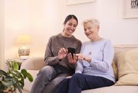 Eine junge Frau und eine ältere Dame sitzen gemeinsam auf einem Sofa. Die jüngere Frau erklärt den Malteser Hausnotruf.