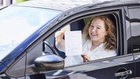Eine junge Frau sitzt hinter dem Lenkrad eines PKW und hält eine Bescheinigung durch das geöffnete Fenster.