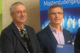 Zwei Männer im Anzug stehen vor einem Banner, einer hält eine Mappe der FAU Universität.
