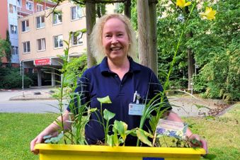 Frau in Arbeitskleidung steht im Grünen und hält eine gelbe Kiste mit Jungpflanzen.