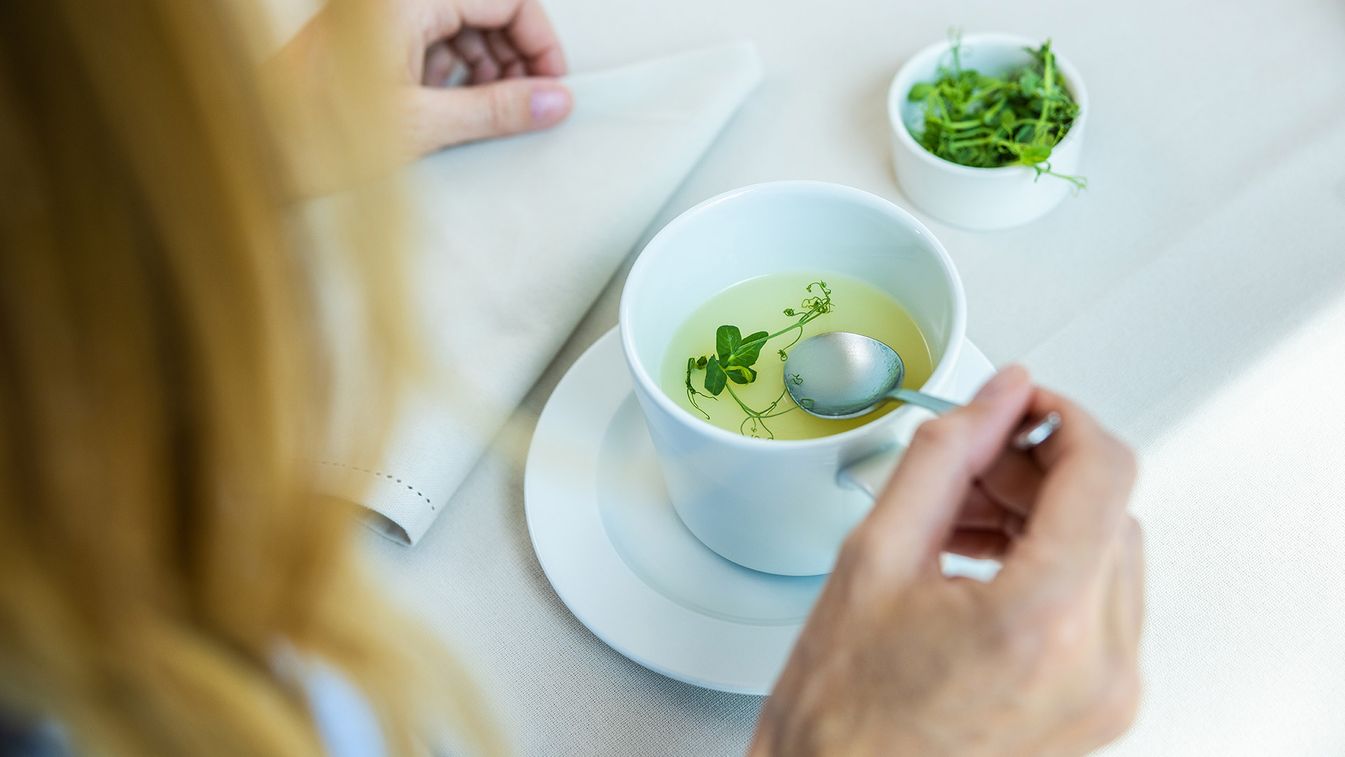 Blick über die Schulter einer Frau auf einen Teller Gemüsesuppe. Die Frau löffelt die Gemüsesuppe.