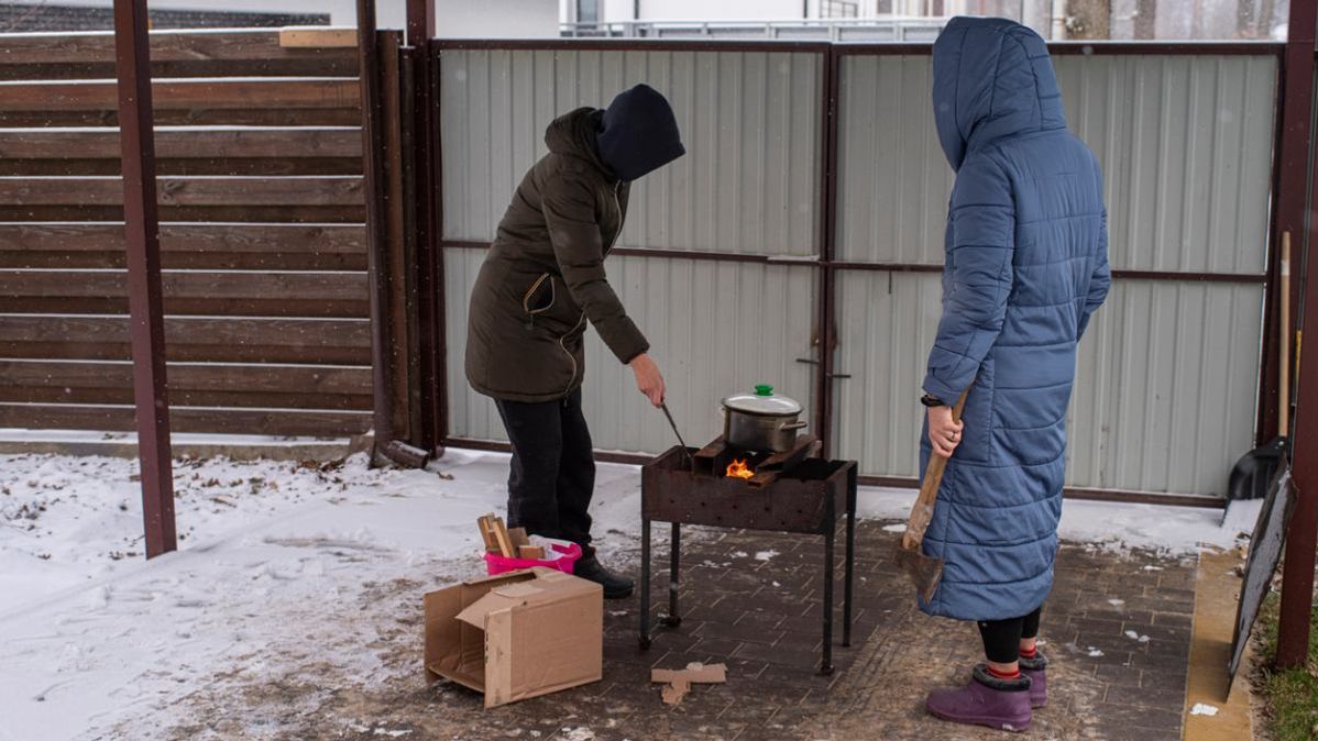 Zwei Menschen in Winterjacken kochen draußen etwas in einem Topf, der auf einem Grill steht.